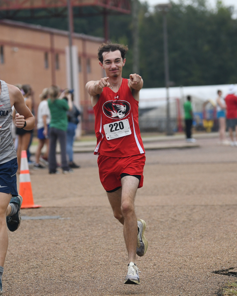 Runner with number 220 in red uniform, pointing forward. Another runner in gray. Background: building, spectators, traffic cone.