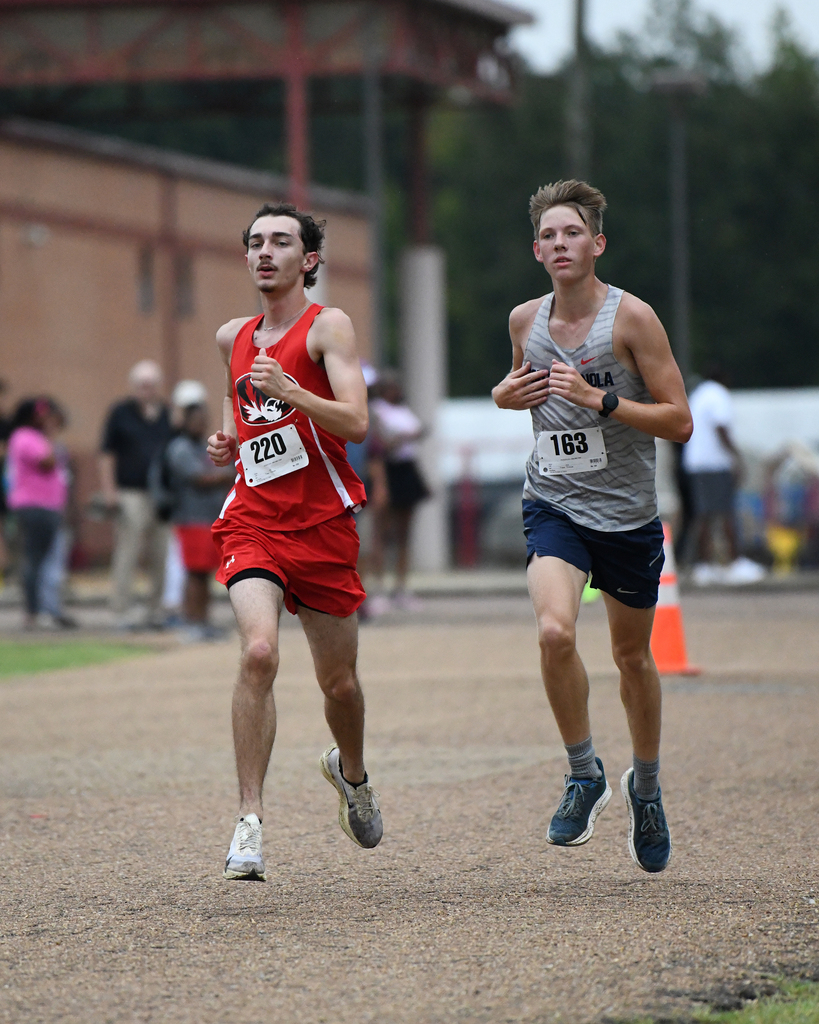Two men in athletic gear run on a dirt track. One wears red, the other gray. Spectators are behind them.