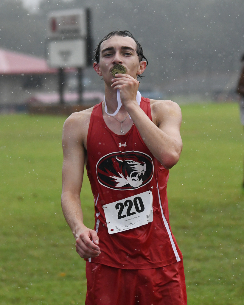 A male athlete in red sports gear, number 220, runs in the rain with a medal in his mouth.