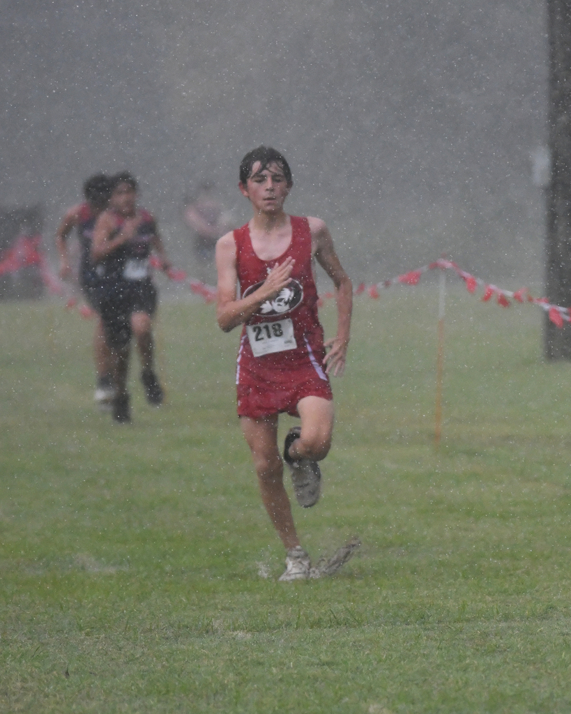 A young runner, number 218, sprints in the rain on a grassy field, surrounded by blurred figures and red ribbon markers.