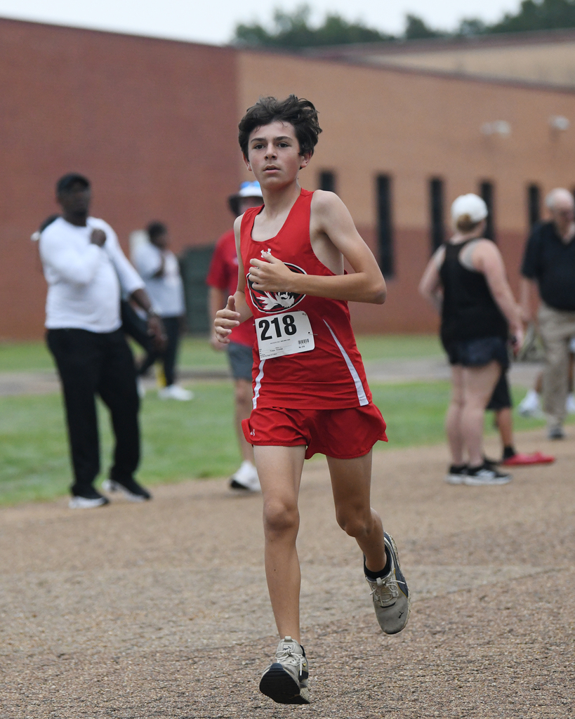A boy wearing a red jersey with the number 218 runs on a gravel track. Spectators stand in the background.
