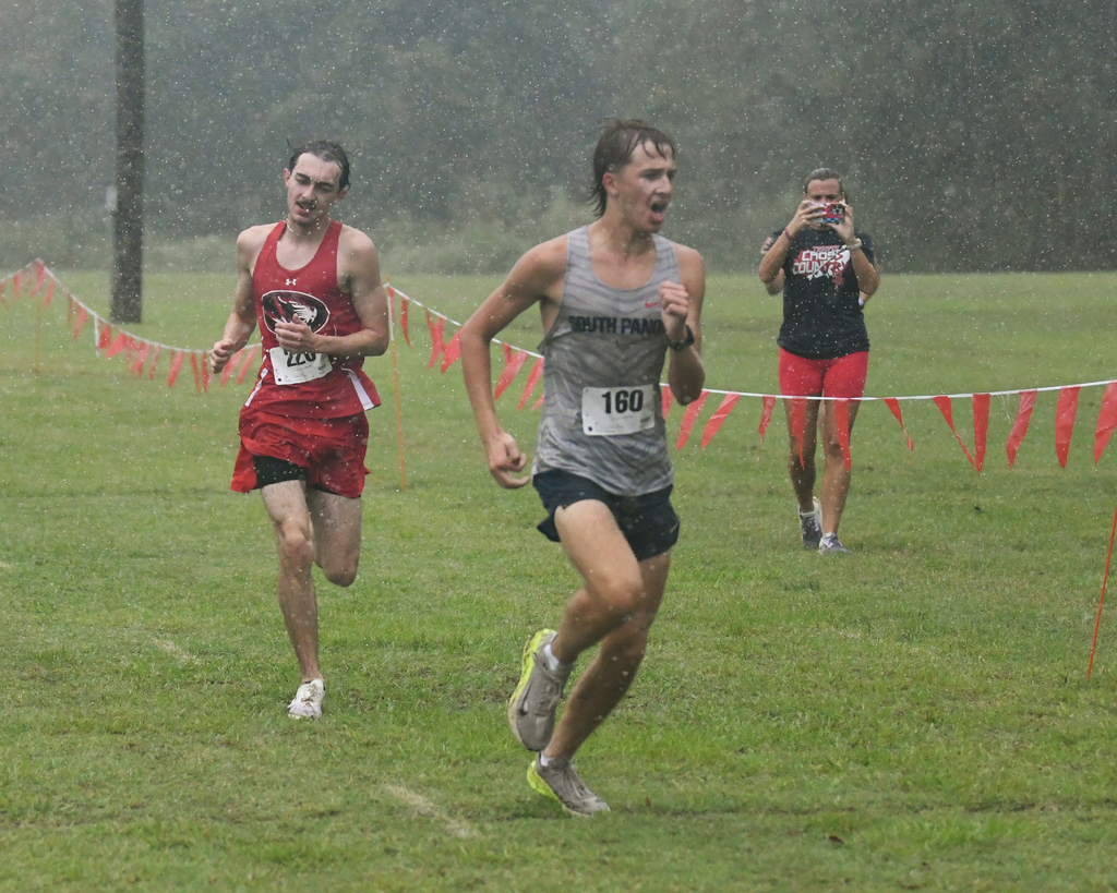 Three people in red and gray sportswear run in a race on grass. One has a camera.