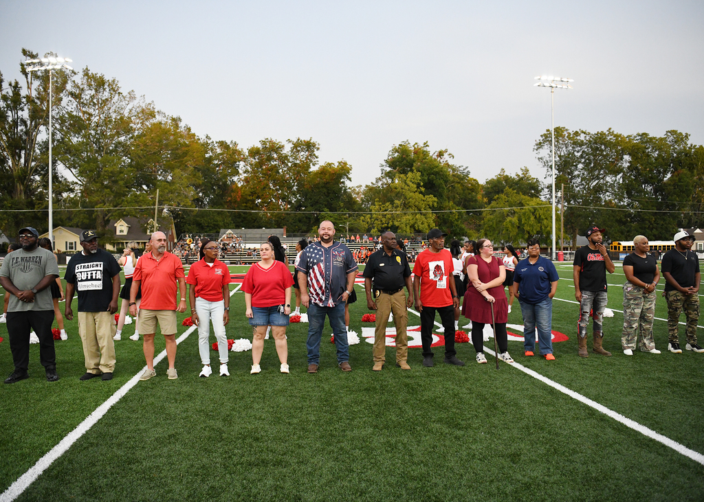 A group of adults in red and white shirts and shorts stands on a football field.