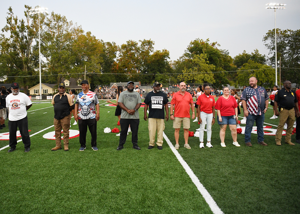 A group of people stands in a line on a football field, dressed in various shirts, including some with patriotic designs.