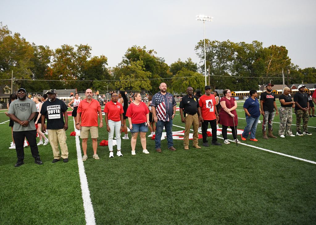 People standing in a line on a football field, with one person wearing a flag. Trees and a building in the background.