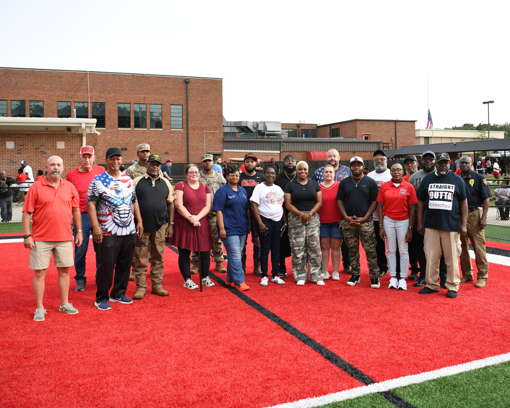 A group of people stand on a red field, wearing diverse clothing. A building with multiple windows is in the background.