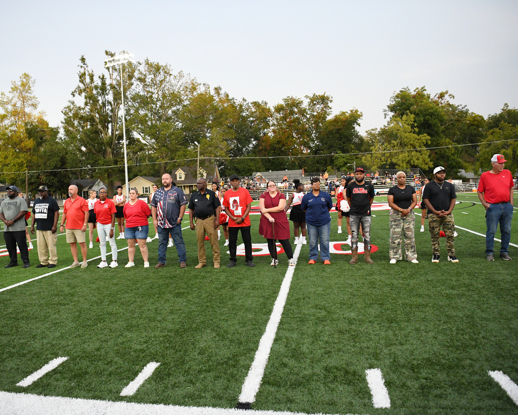 A group of people in various clothing stand on a football field, lined up with white lines.