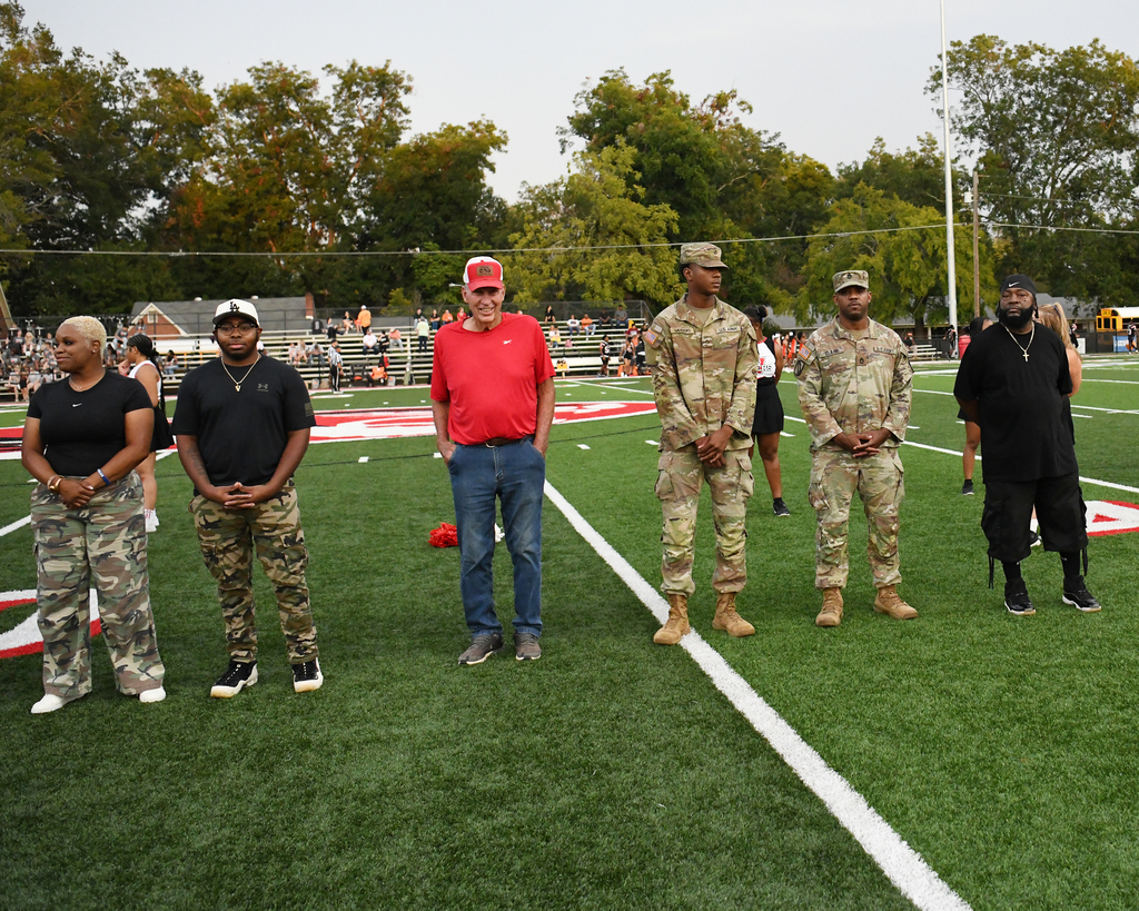 A group of people stand on a football field with white lines. Trees are in the background.