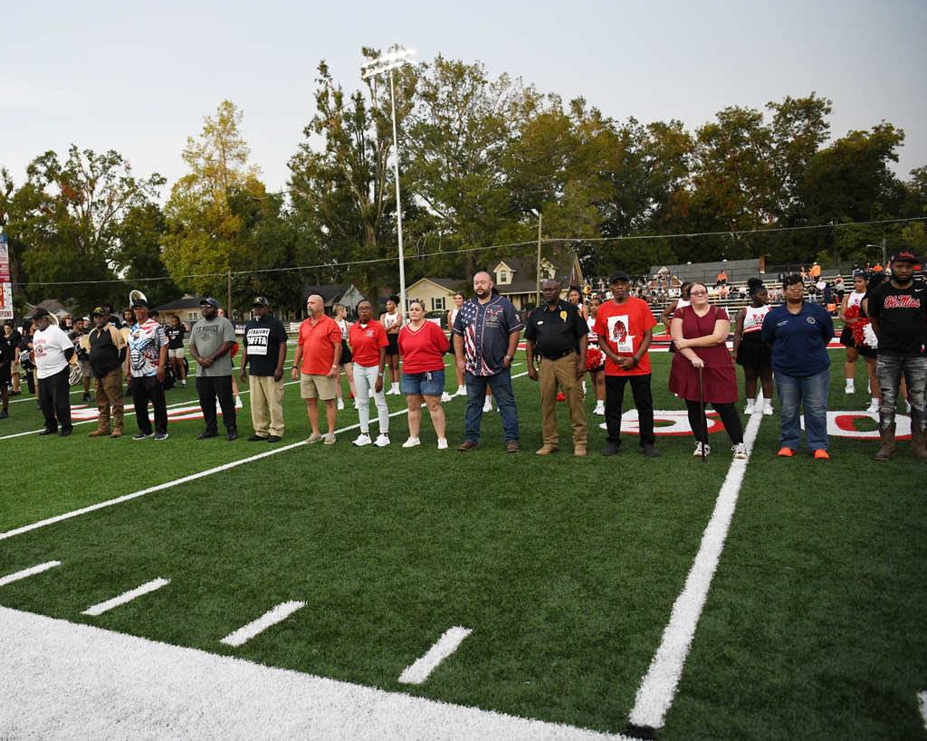Group of people standing on a football field, facing forward. Trees and buildings in the background.