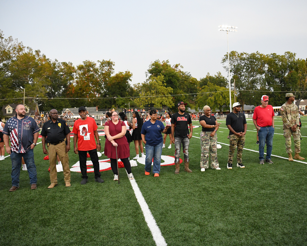 A group of people stands on a football field with a white line. Some wear hats and sneakers. Trees are visible in the background.