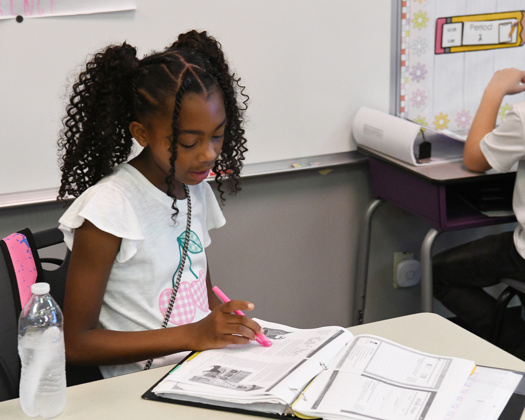 A girl in a classroom writes in a book, using a pink marker. Behind her, a desk holds a water bottle.