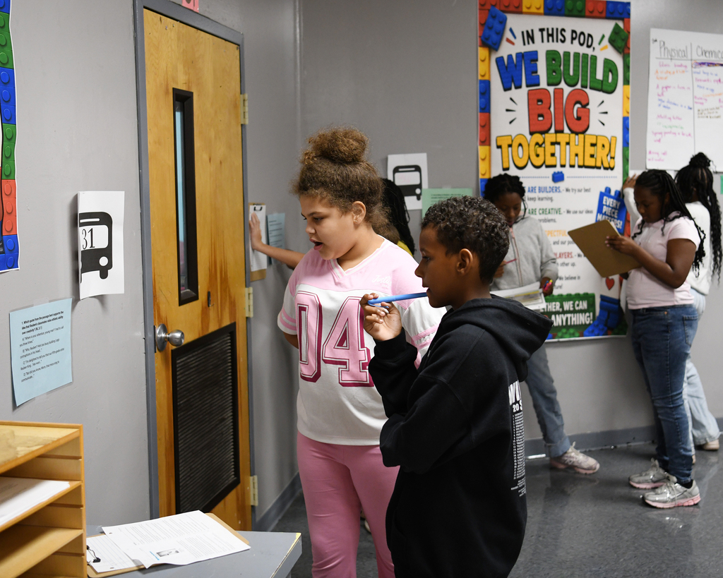 A classroom with a sign "We Build Big Together". Students, one holding a clipboard, stand in front of a bulletin board.