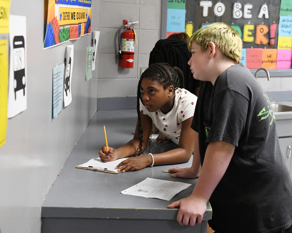 A classroom with a student writing in a notebook and another student reading over her shoulder.