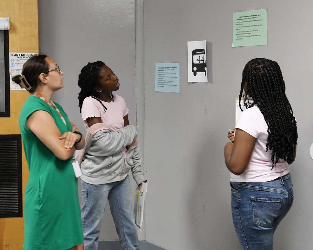 Three women face a gray wall with a bus route sign. The left woman wears glasses, the middle one a pink shirt, and the right one a white shirt.