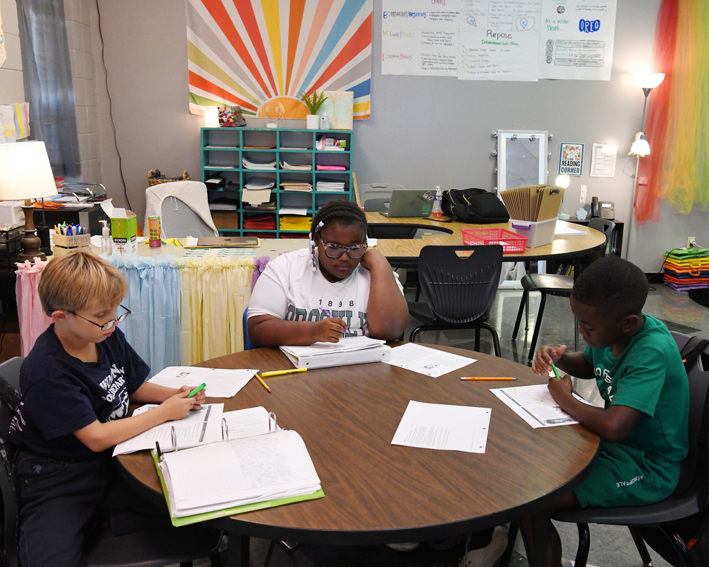 Three children sit at a table with papers and pens, one in a white shirt, another in a green shirt, and the third in a black shirt.