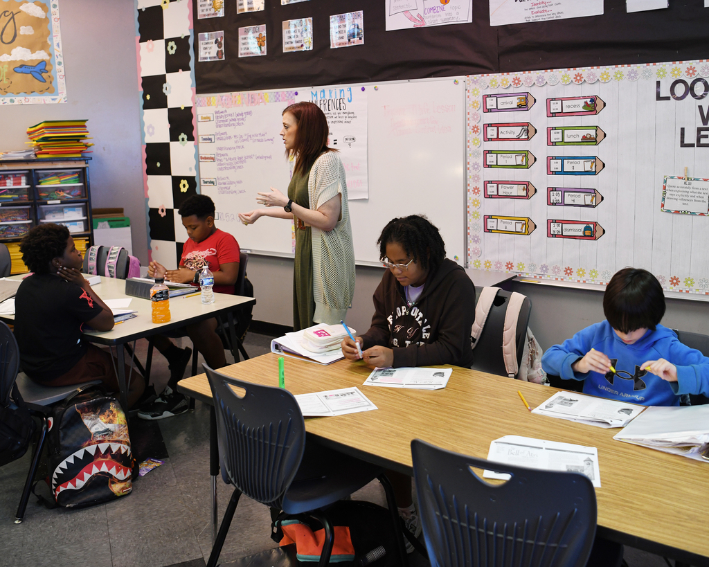 Woman standing at front of classroom teaching students. Students sitting at desks, some with papers and pens.