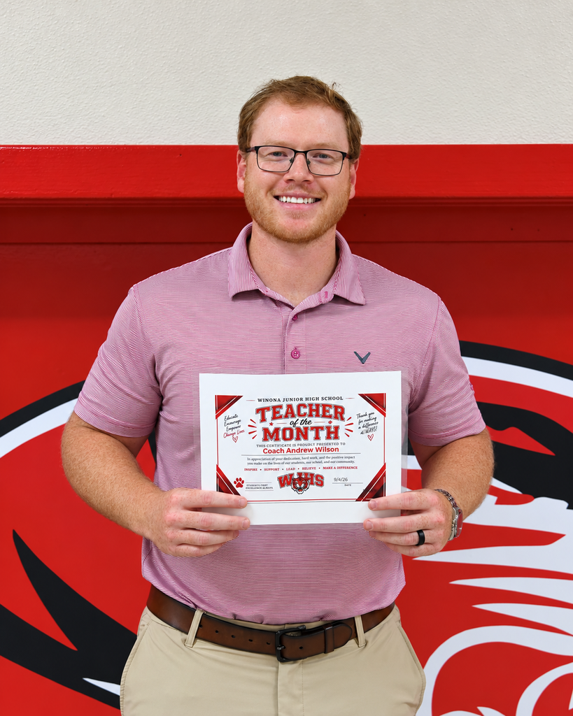 A man holds a certificate that says Teacher of the Month. He is smiling. The wall behind him has a red and black design.