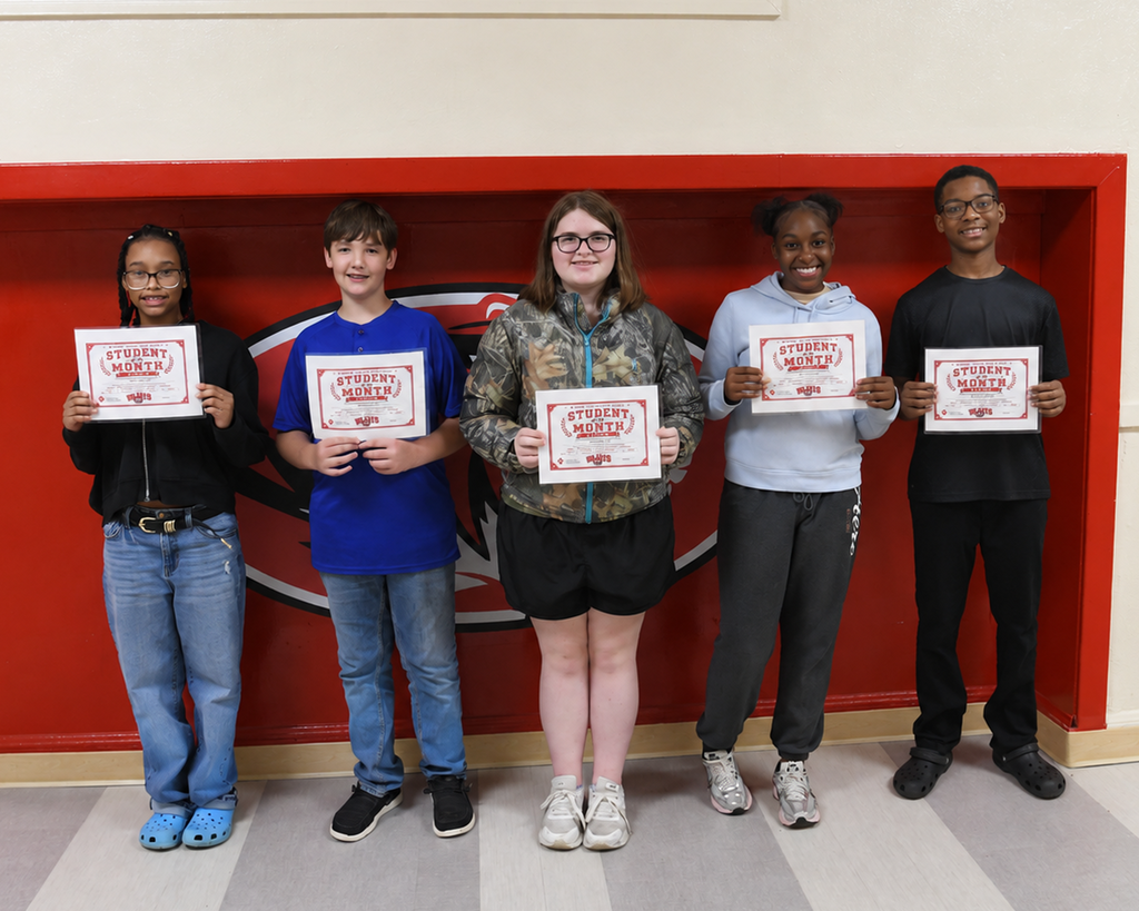 Five people stand in front of a red wall with a large logo, holding certificates. They wear casual clothing.