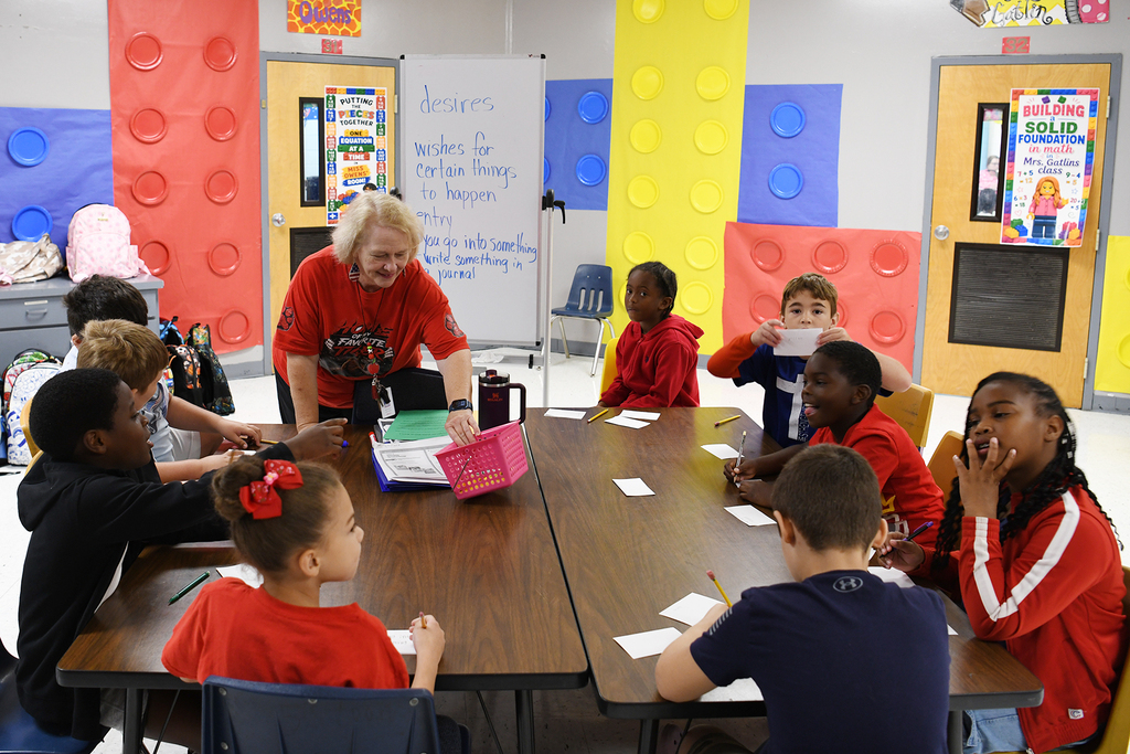 Classroom setting with a woman standing at a table and children seated at a long table.