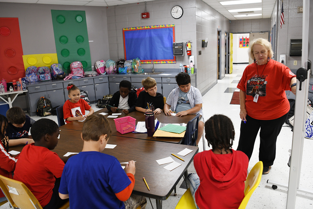 A classroom with students sitting around a table, a teacher standing nearby. Walls have colorful posters and a clock.