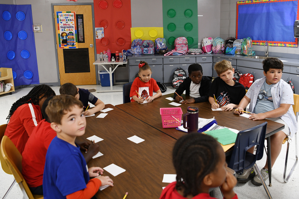 A classroom with a group of children sitting around a table. One child writes on paper.