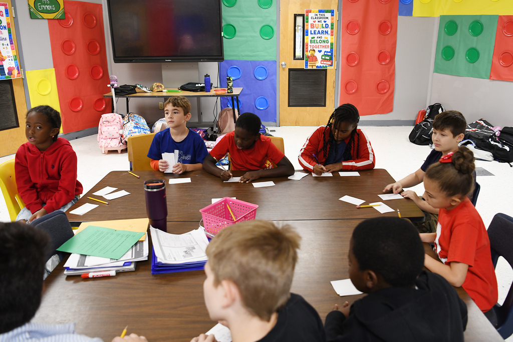 Children in a classroom with Lego wall blocks, seated around a table. Some have papers, pencils, and colored folders.