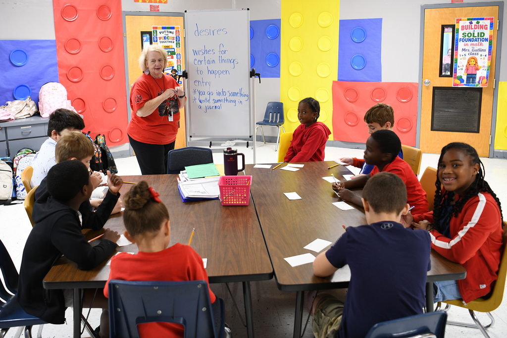 Children seated at a table, looking at a teacher who holds a microphone and a marker in a colorful classroom.