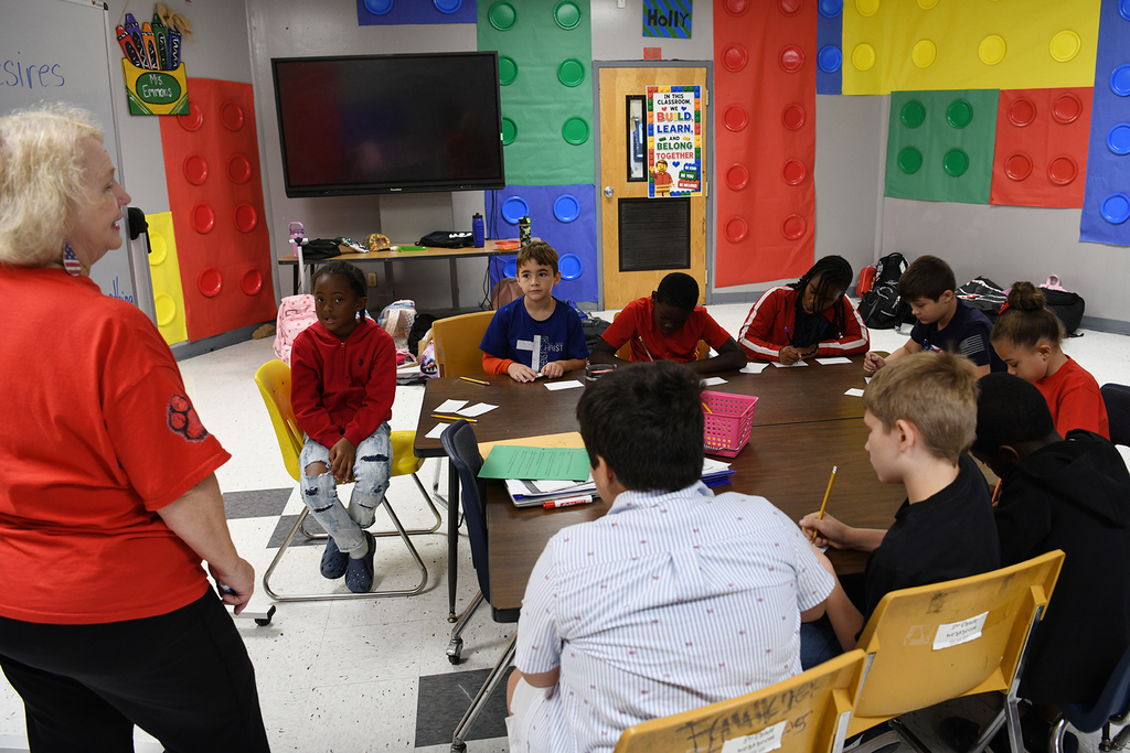 An adult stands before a group of students around a table in a colorful classroom.