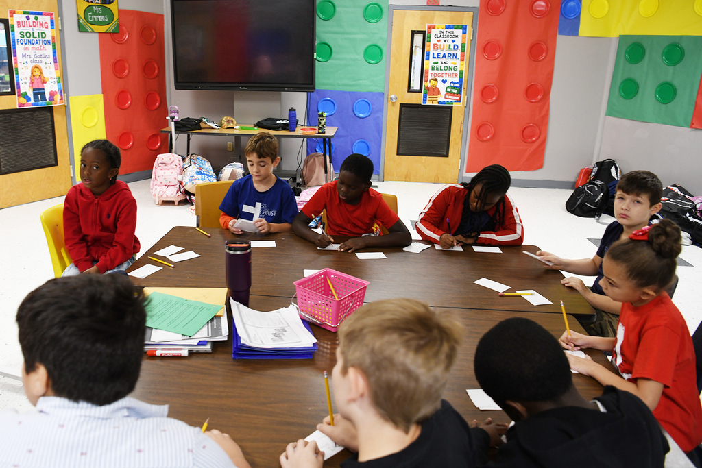 A classroom with young students seated around a table, working on papers. Brightly colored blocks adorn the walls.