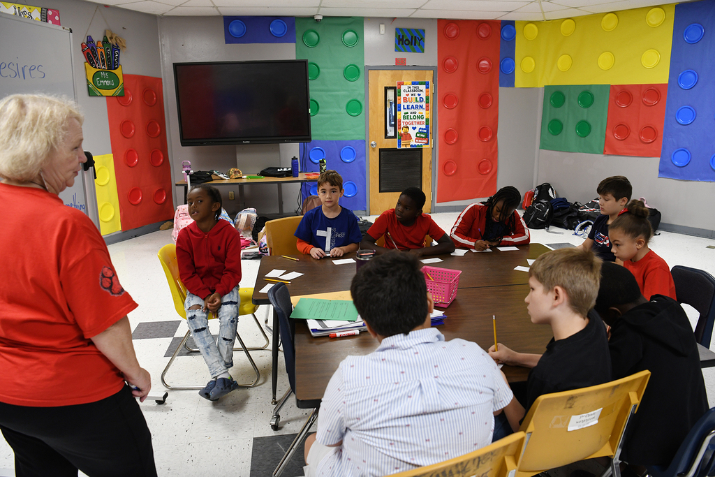 A woman stands in front of a table with young kids in a classroom with colorful Lego walls.