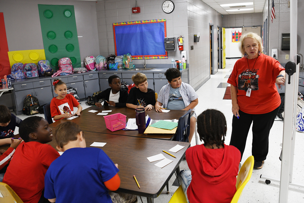 A woman leads a group of children seated at tables in a classroom, with colorful decor on the walls.