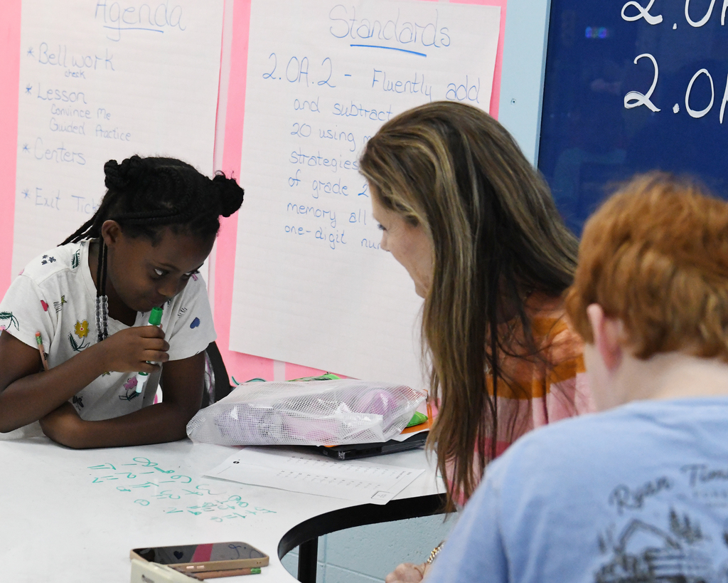 A classroom with two students and a teacher; a girl writes on a whiteboard while the teacher and boy listen.