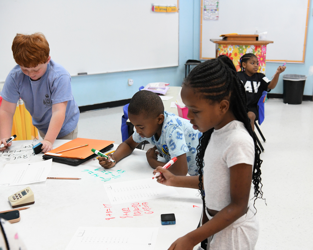 Children in a classroom work on a large paper. One child writes in red marker. A whiteboard is on the wall.
