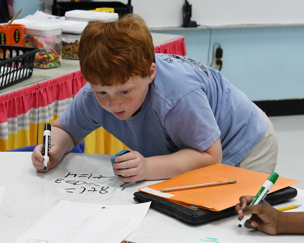 A child writes on a desk while another holds a marker. Behind them, shelves with items are visible.