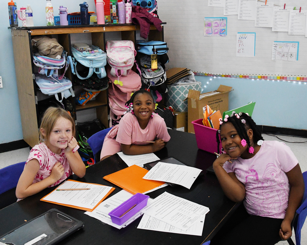 Three girls seated at a table, engaged in studying, with school supplies and bags in the background.