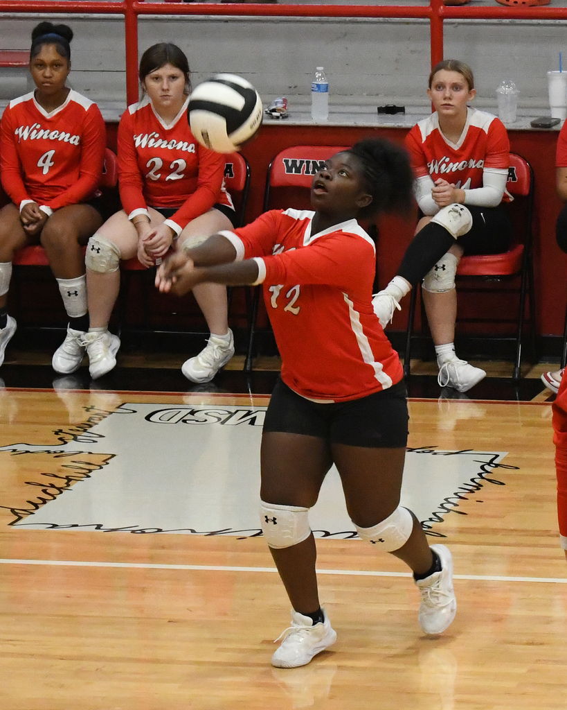An indoor volleyball player in red uniform hits a ball with knees bent. Behind are teammates in red uniforms.