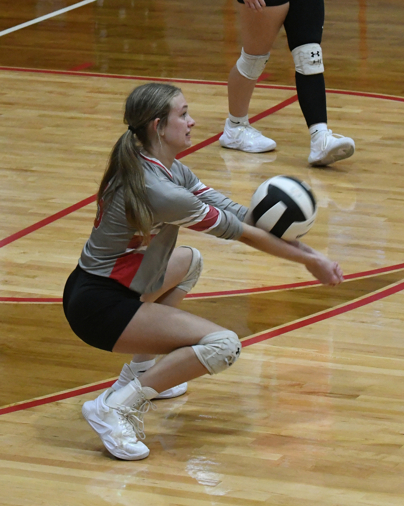 A female volleyball player in a gray and red uniform crouches on a court to catch a ball.