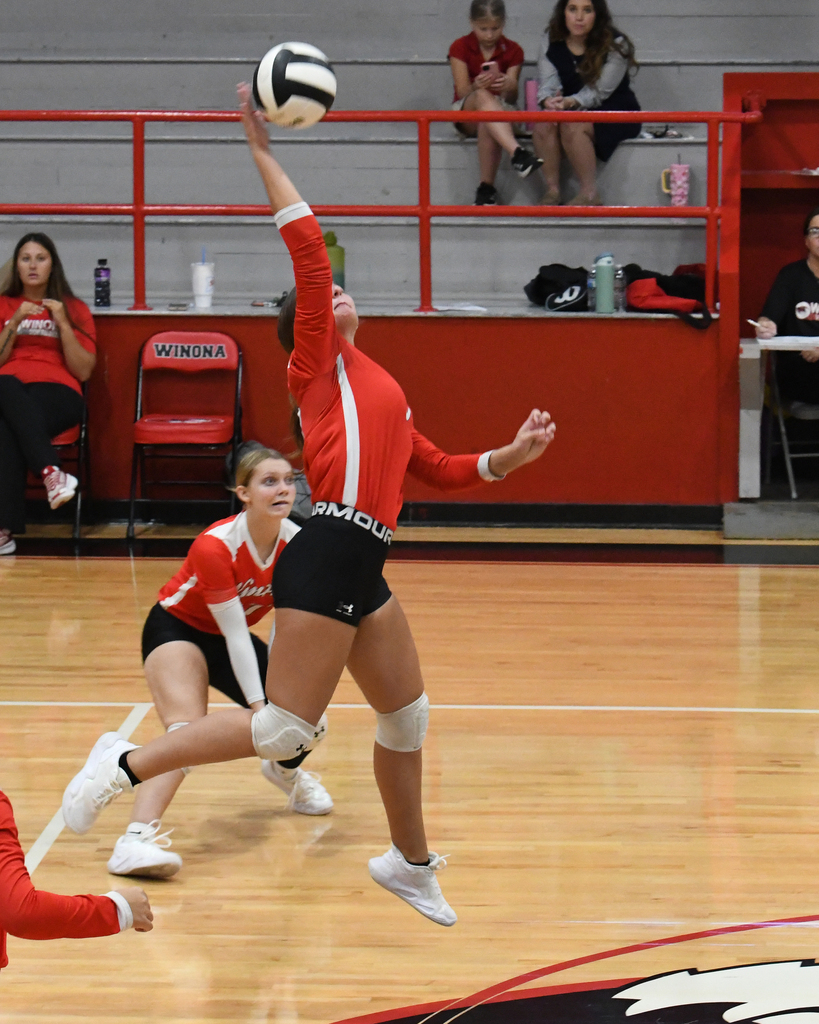 A female volleyball player in mid-air, jumping to spike the ball. Behind, two teammates on a wooden court. Spectators sit on red bleachers.