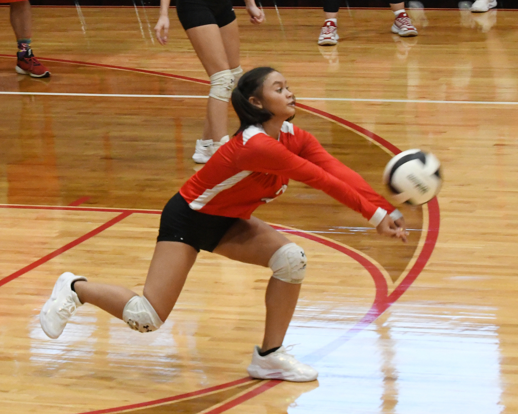 A female volleyball player in red and black uniform lunges, extending arms to hit a ball. Behind her, others on a court with red lines.