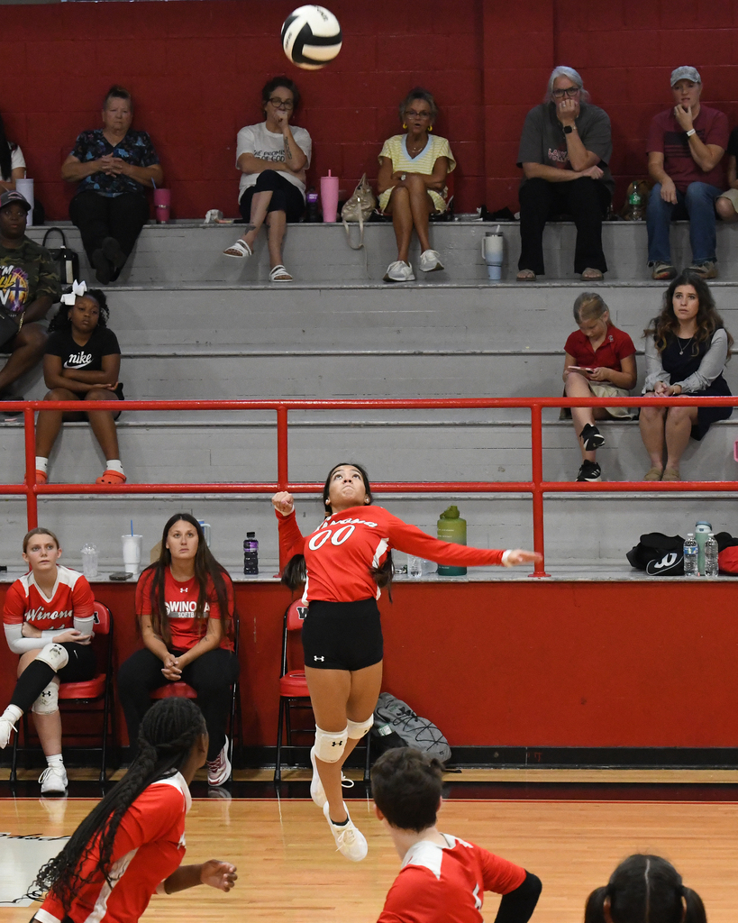 A volleyball player jumps to hit a ball in a gym. Spectators sit on red bleachers.