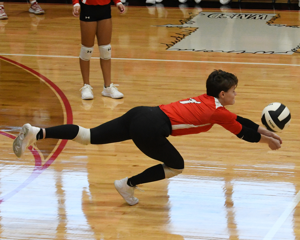 A young person in red and black playing volleyball, diving to touch the ball. A teammate stands behind.