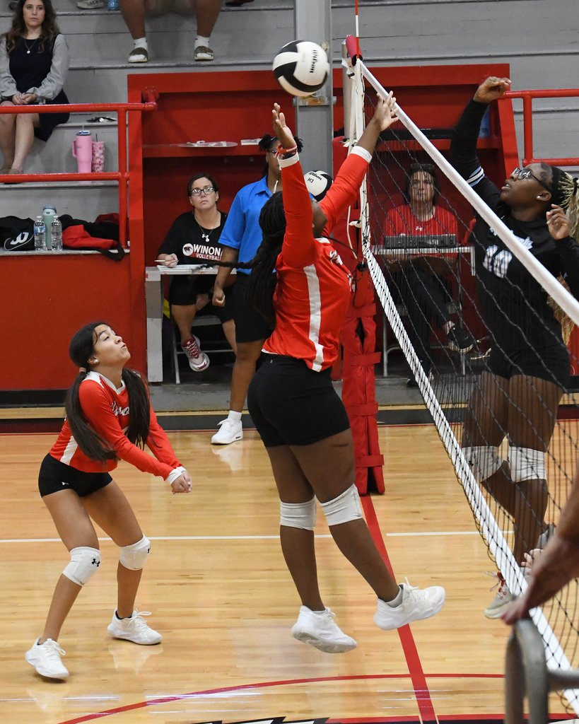 Players in red and black uniforms playing volleyball on a court with a net. Two players block an opponent.