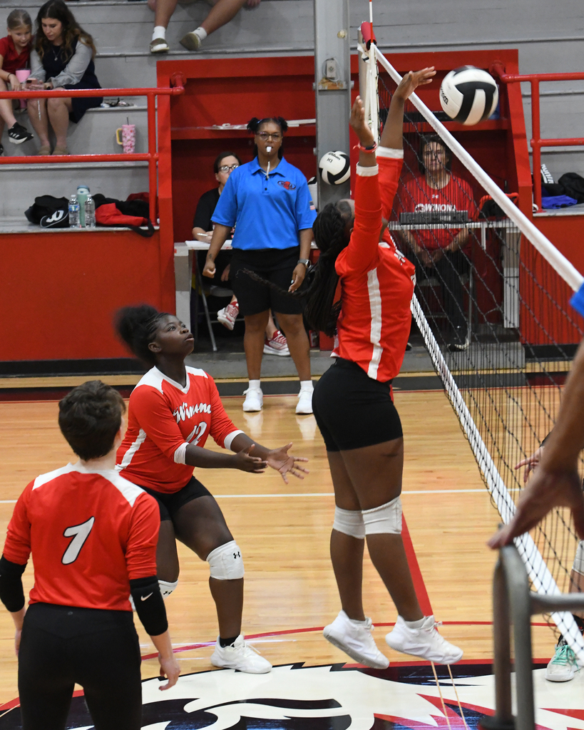 Players in red uniforms, with one jumping to block the ball, on a volleyball court.