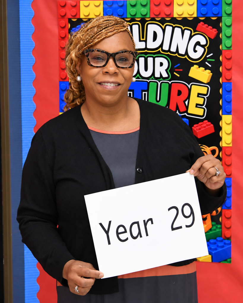 Woman wearing glasses and a black cardigan holds a white sign reading "Year 29" in front of a backdrop with a colorful Lego pattern.