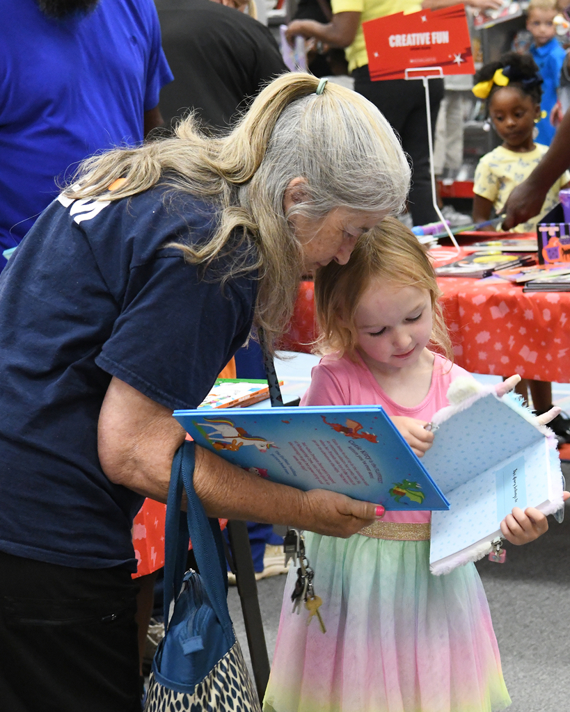 Woman with long gray hair bends over to read a book with a smiling child in a pink dress.