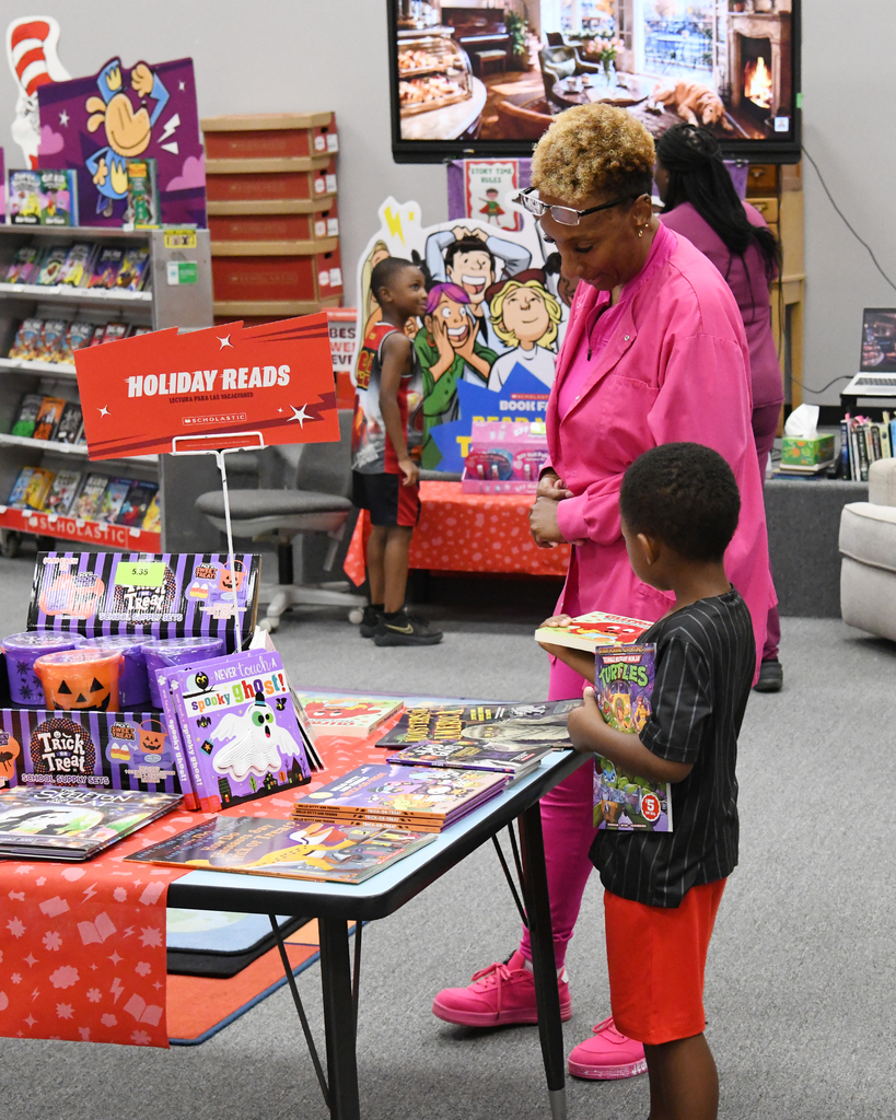 An adult woman and a child browse a table of books with a holiday reads sign in a room.