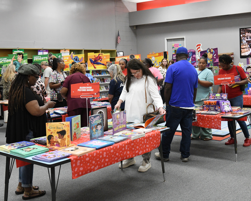 People at a book fair, browsing various books on tables covered with red cloth. Background includes bookshelves and a bulletin board.
