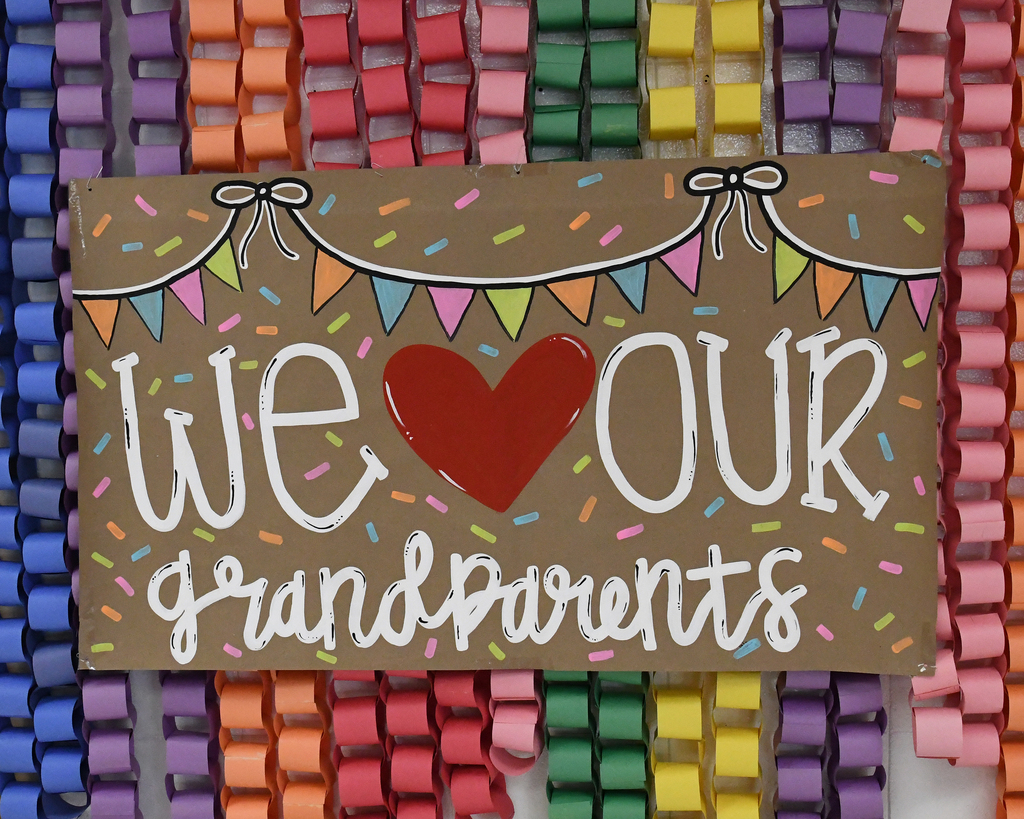 A cardboard sign with white letters reading "We love our grandparents" and a red heart. It is adorned with colorful paper banners and confetti.