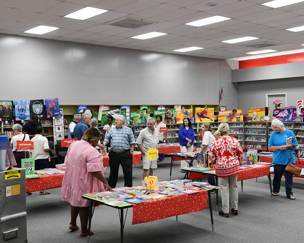 People browsing at tables filled with books in a large, well-lit room with bookshelves in the background.