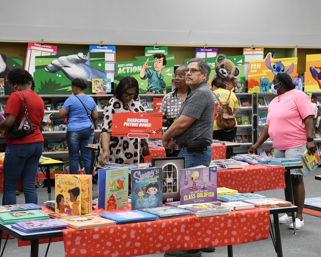 People at a book fair with tables displaying children's books and colorful posters on the wall.